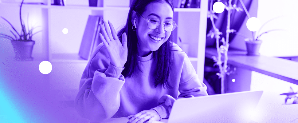 Cheerful woman video calling, waving at the laptop screen in a monochromatic blue home office setting